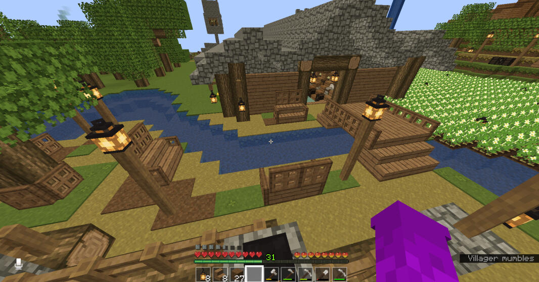 An image of a small area behind the villager trading hall, decorated with benches, lamp posts, and a small river connected to a pond. The trading hall is built almost entirely out of wood, with a cobblestone roof. To the far right side of the image, you ca